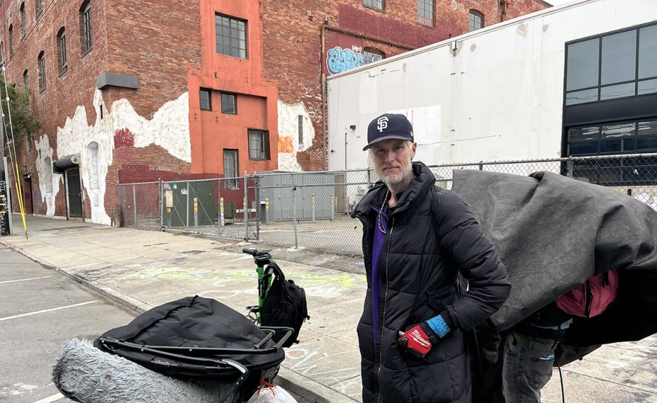Man in a black jacket and cap pushing a cart covered with a tarp, standing on an urban street with red brick building and graffiti in the background.