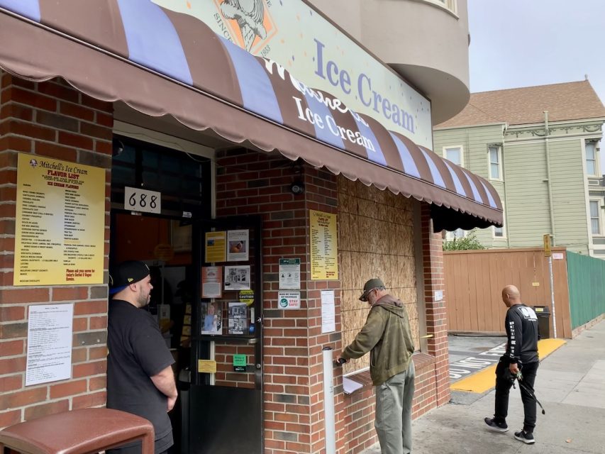 Customers stand outside an ice cream shop beneath an awning that reads "Ice Cream." One person reads a posted menu while another person looks at items on a counter beside a boarded-up window.