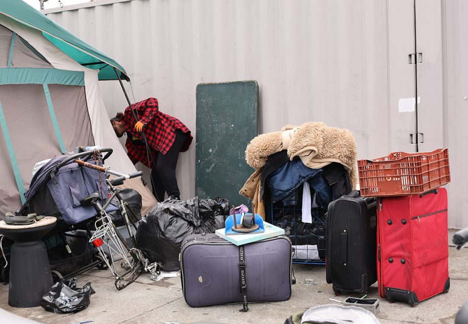 A person in a red plaid shirt peers into a tent surrounded by various bags, a stroller, luggage, and piled belongings, adjacent to a corrugated metal wall that seems to sweep across the scene.