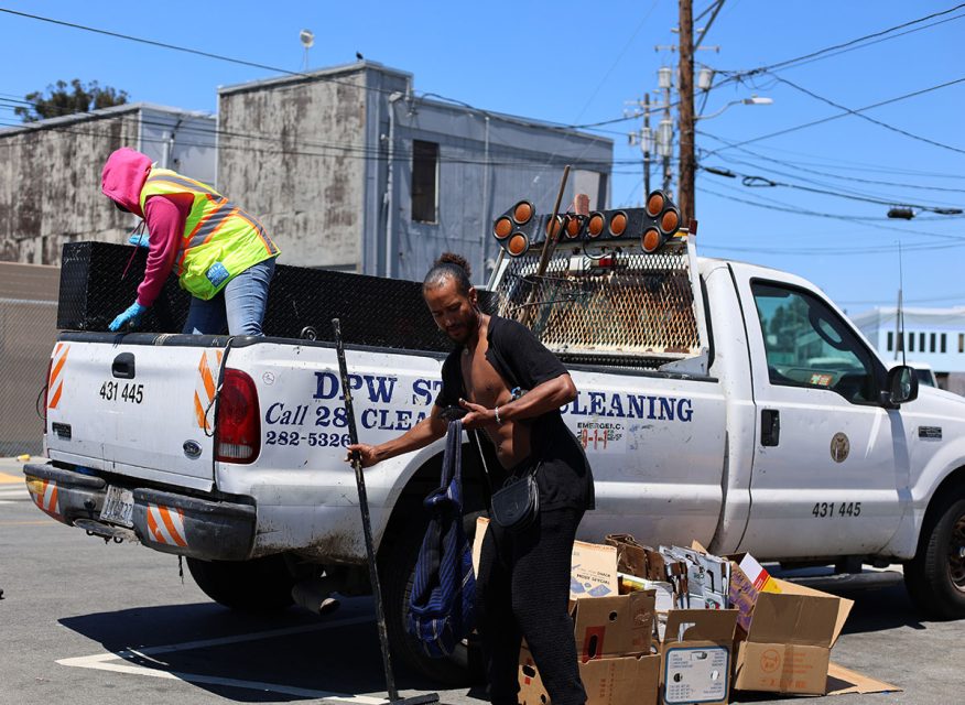 A person in a yellow vest stands in the bed of a DPW truck, while another person with a broom and bags is nearby, ready to sweep up the cardboard boxes scattered on the ground.