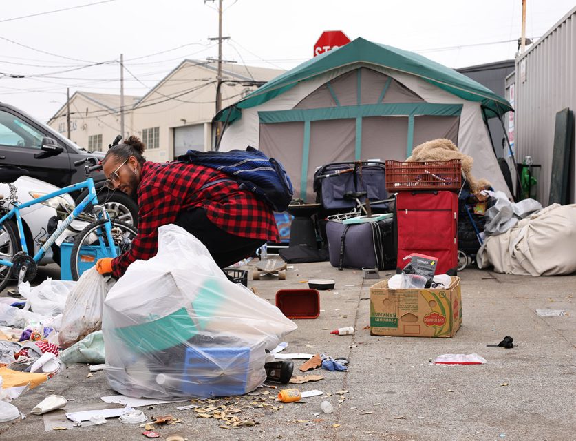 A person in a red plaid shirt and glasses is sweeping up trash in front of a tent with various items like luggage, boxes, and bicycles scattered around.