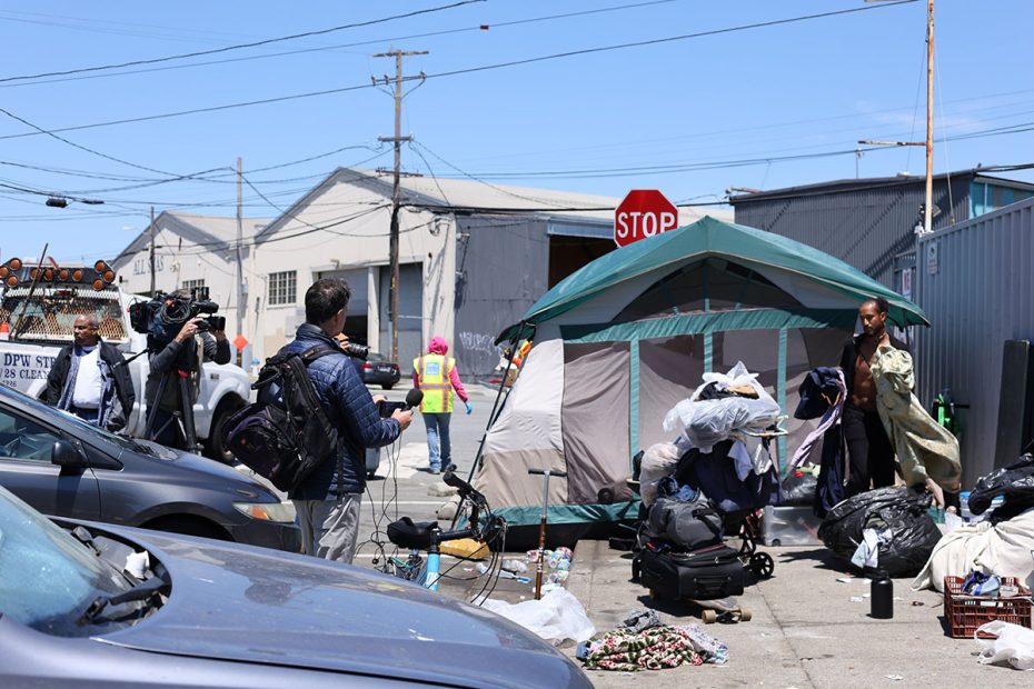 People are gathered near a tent surrounded by belongings on a city street, as a news crew covers the scene. Utility workers are present, preparing for the scheduled sweep.