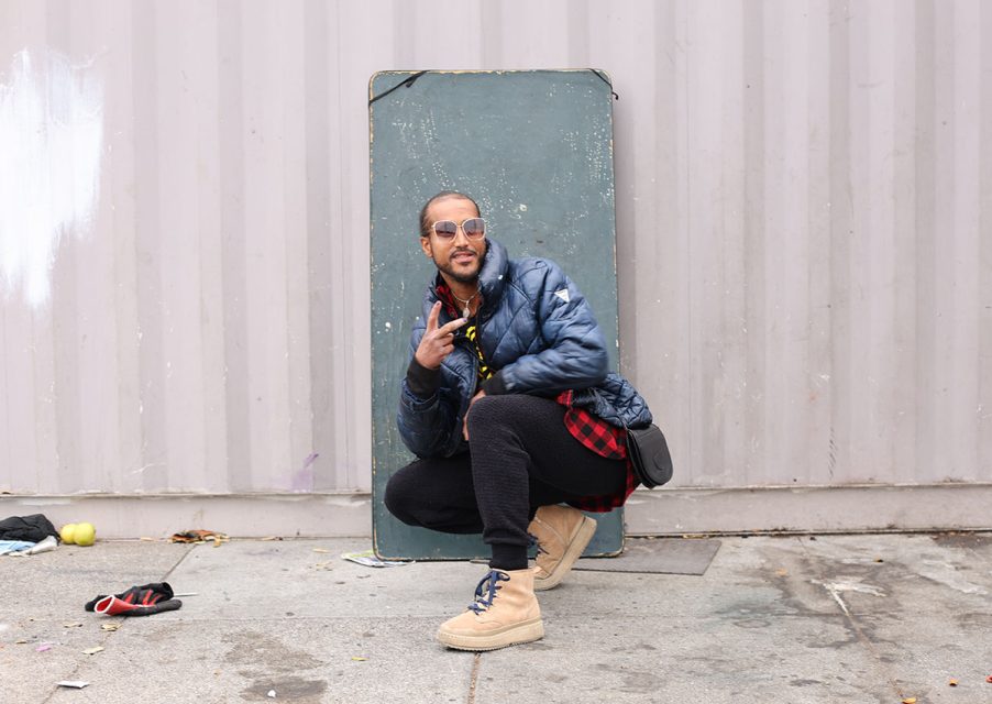 A person wearing sunglasses, a dark puffer jacket, and beige boots squats in front of a grey partition, making a peace sign gesture with their hand. Various small items are scattered on the ground, as if caught in a sweeping moment.