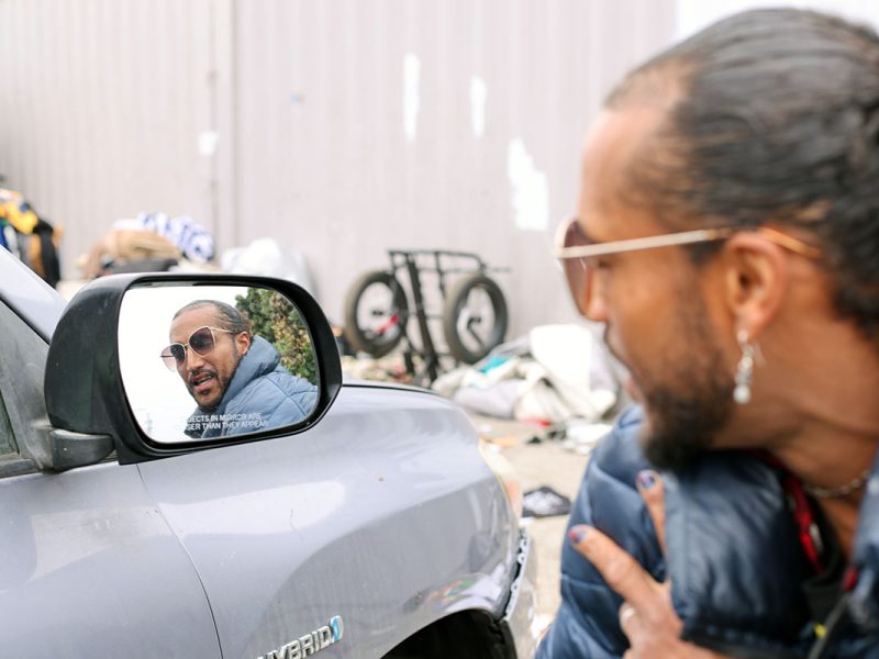 A person is reflected in the side mirror of a vehicle, looking at themselves. They wear sunglasses and have their hair tied back in a neat sweep. The background shows miscellaneous items and a bicycle.