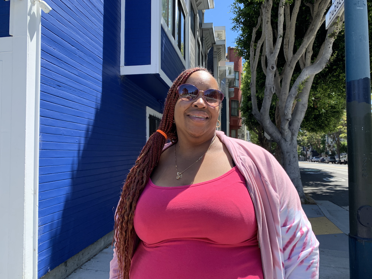 A woman with long braids and wearing sunglasses and a pink top stands smiling on a sunny day in front of a blue building with a tree in the background.