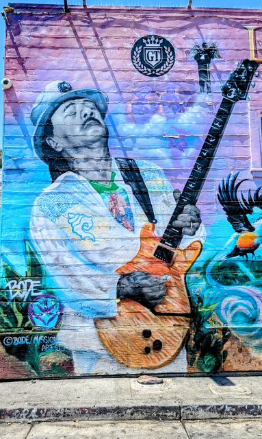 A colorful mural on a wall depicts a musician with a guitar. The artist's name and details are visible on the bottom left corner.