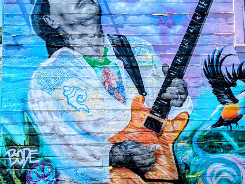 A colorful mural on a wall depicts a musician with a guitar. The artist's name and details are visible on the bottom left corner.
