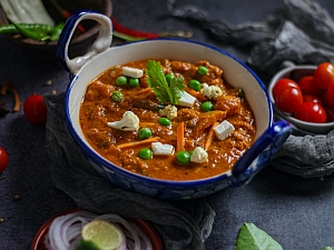 A bowl of curry filled with mixed vegetables, garnished with a sprig of cilantro, surrounded by cherry tomatoes, sliced onion, and a lime wedge.