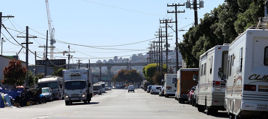 A street lined with parked RVs and utility poles. A few trees and buildings are visible in the background.