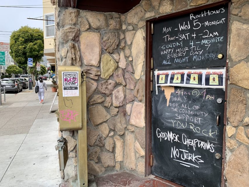 Chalkboard sign on a stone wall lists bar hours and rules. Nearby, a painted metal box with stickers stands on a sidewalk with cars and pedestrians.