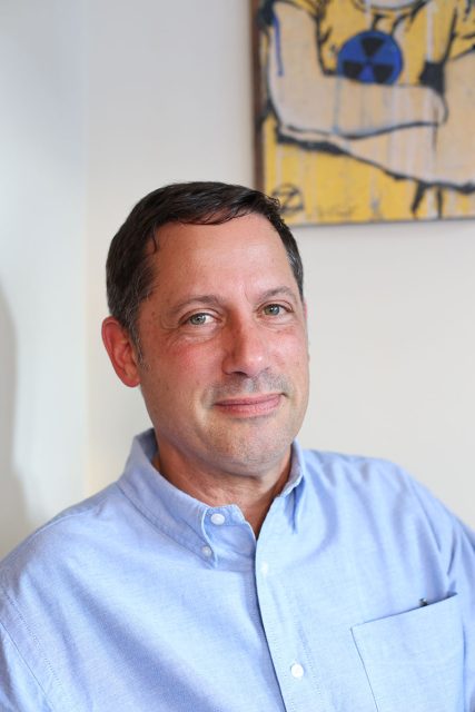 A man with short dark hair wearing a light blue button-up shirt sits in front of a white wall with a painting featuring abstract art in the background.