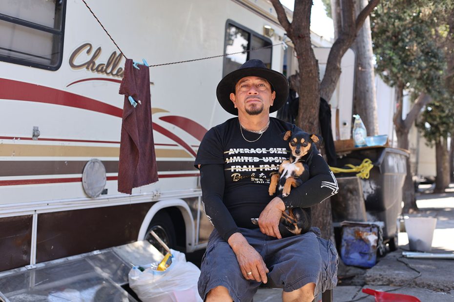 A man wearing a hat and casual clothing sits outside, holding a small dog. Behind him, there's a trailer and some personal belongings scattered across the ground as if swept by the wind.