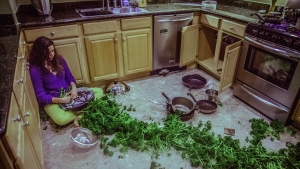 A woman sits on the kitchen floor with her back against a cabinet. Pots, pans, and green leafy vegetables are scattered across the floor. The kitchen appears disorganized.