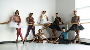 Seven ballet dancers in various poses, wearing different styles of ballet attire, stand and lean against a ballet barre in a well-lit studio.