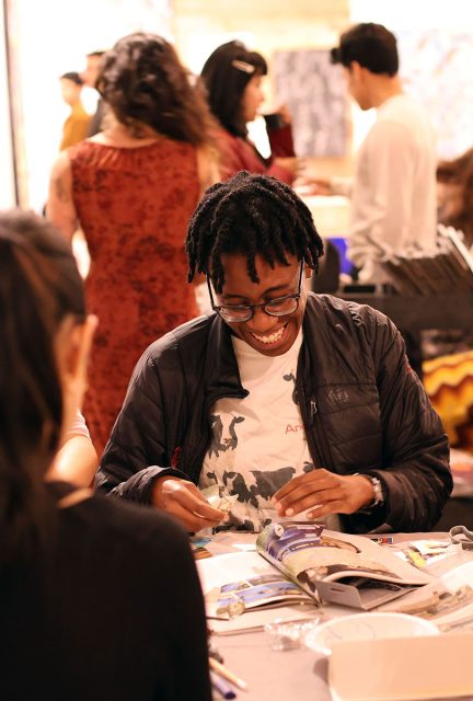 Person wearing glasses and a jacket smiles while working on an art project at a table. Other people are engaged in activities in the background.