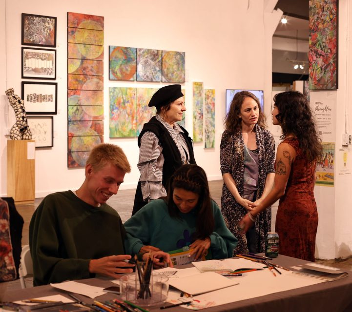 A group of people is interacting in an art gallery. Two people are seated at a table sketching, while three others are standing nearby and conversing. Various artworks are displayed on the walls.