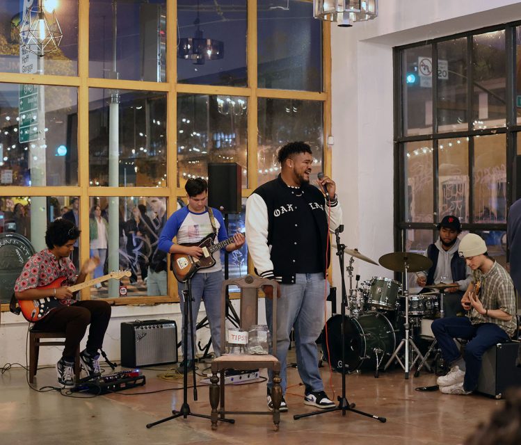 A band performs in a cozy, modern setting. The lead singer is at the microphone, while the guitarist, bassist, keyboard player, and drummer play their instruments. The room has large windows and hanging lights.