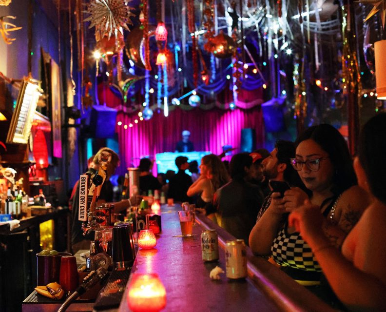 A vibrant bar scene with colorful lights and decorations. People are gathered around the counter, with a DJ performing in the background. Drinks and candles are on the bar.