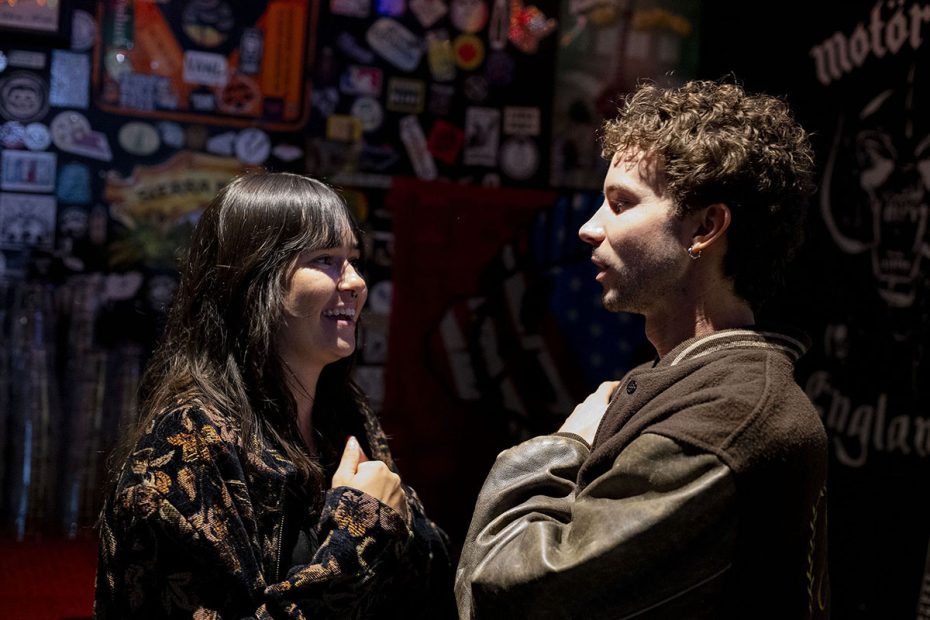 Two people stand facing each other in a dimly lit room decorated with various stickers and posters. Both are smiling, engaging in conversation.