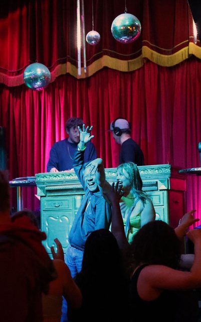 People dance in a club with a red curtain backdrop, two DJs perform behind a wooden console, and disco balls hang from the ceiling.
