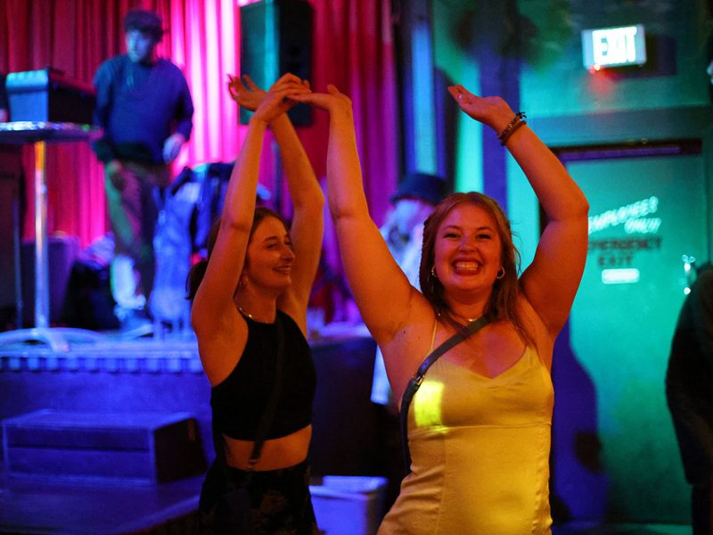 Two women are dancing with raised arms in a dimly lit venue with red curtains in the background. The woman on the right is wearing a yellow dress and smiling broadly.