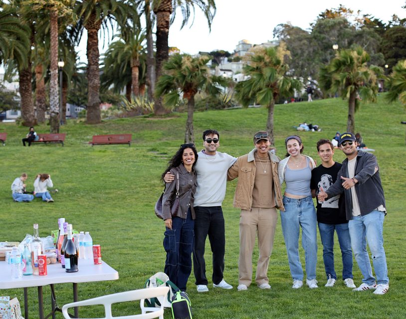 Six people are standing in a park, smiling for a group photo. A table with drinks and food is on the left. Palm trees and other park visitors are visible in the background.