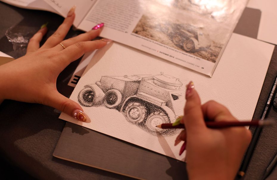 A person is drawing a detailed sketch of a vehicle, using a reference book with an image of the same vehicle. The person's nails are painted vibrantly.