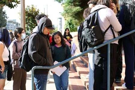 A group of students, some holding papers, stand in line on outdoor steps. Many have backpacks, and some are wearing hoodies. The setting appears to be a school entrance.