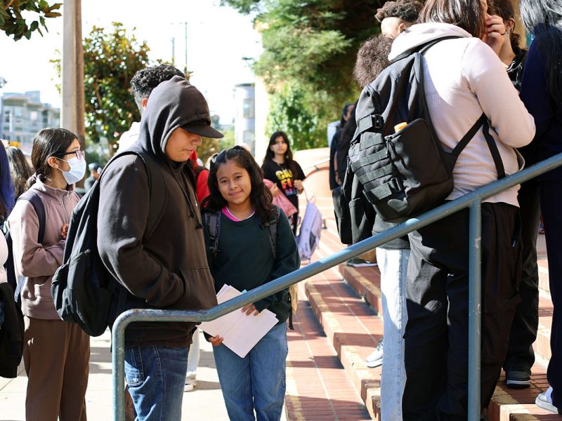 A group of students, some holding papers, stand in line on outdoor steps. Many have backpacks, and some are wearing hoodies. The setting appears to be a school entrance.
