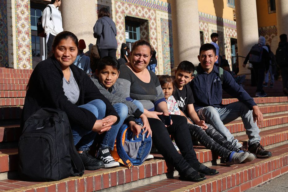 A group of six people, including two women, two men, and two children, sit on steps in front of a building with decorative tiles and columns.