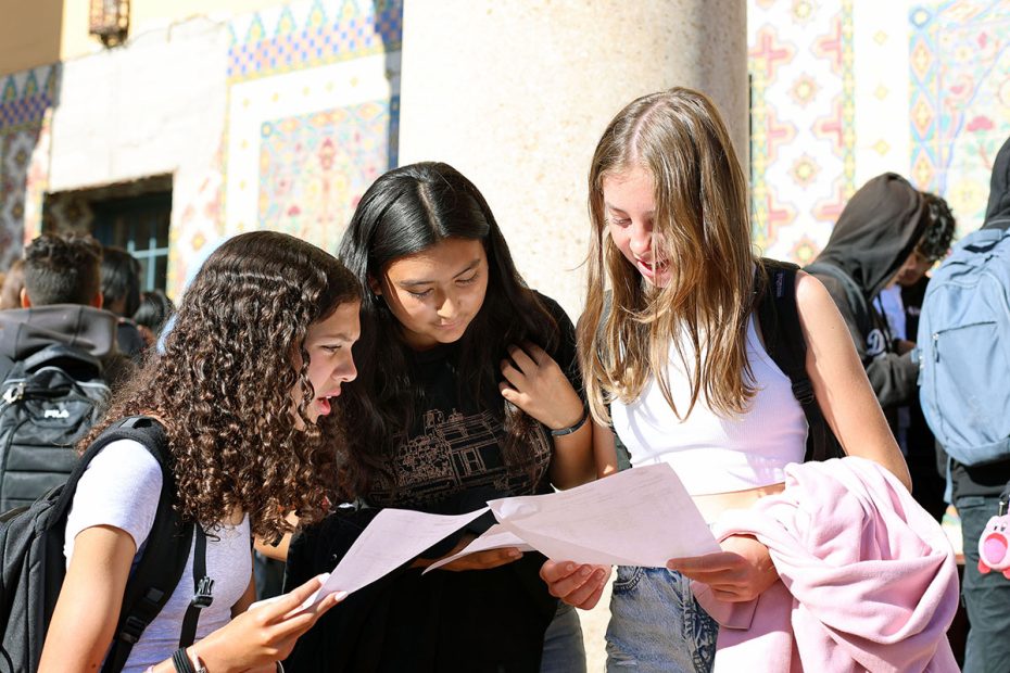Three teenage girls stand outdoors looking at papers, appearing engaged and focused. Behind them, other students and a building with colorful tiles are visible.