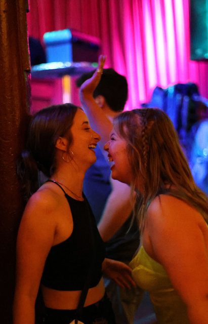 Two people are smiling and laughing together near a wall in a dimly lit venue with pink and purple lighting in the background. A third person is visible in the background raising a hand.