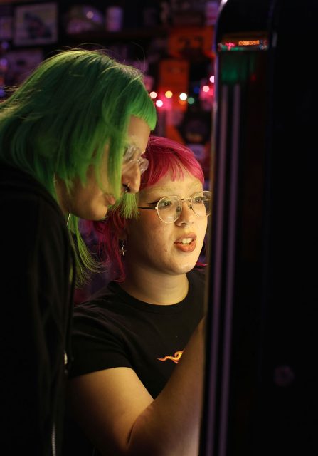 Two individuals with brightly colored hair, one green and one pink, playing an arcade game together in a dimly lit setting.