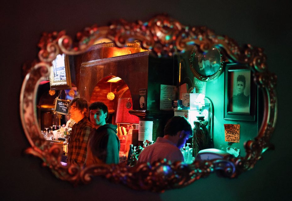 A decorative mirror on a wall reflects three people in a dimly lit bar, with colorful lighting and various posters on the wall.