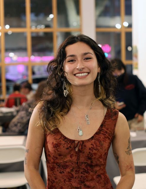 A person with long wavy hair smiles at the camera, wearing a red patterned sleeveless top, silver earrings, and layered necklaces. There is a seated crowd in a room with large windows in the background.