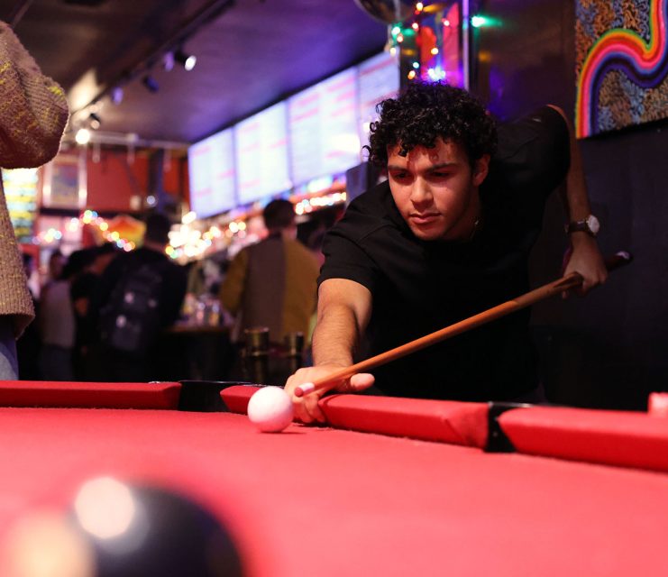 Person aiming a pool cue at a white ball on a red pool table in a dimly lit bar with colorful decorations and people in the background.