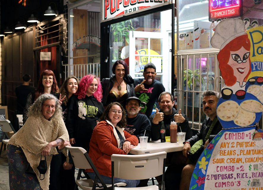 A group of people, some standing and some sitting at a table, pose for a photo outside a brightly lit pupusa restaurant at night. A colorful signboard displays menu items.