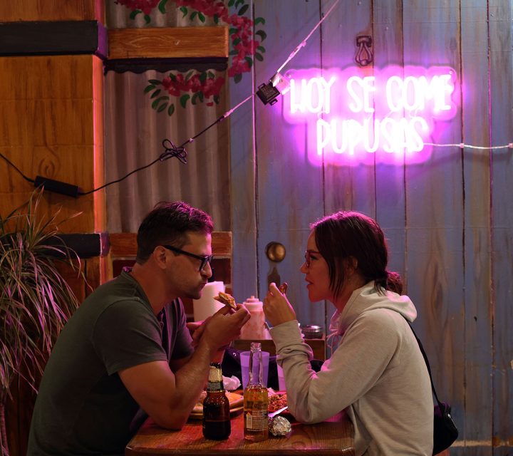 A man and woman sit across from each other at a small wooden table, eating and talking under a neon sign that reads "Hoy Se Come Pupusas.
