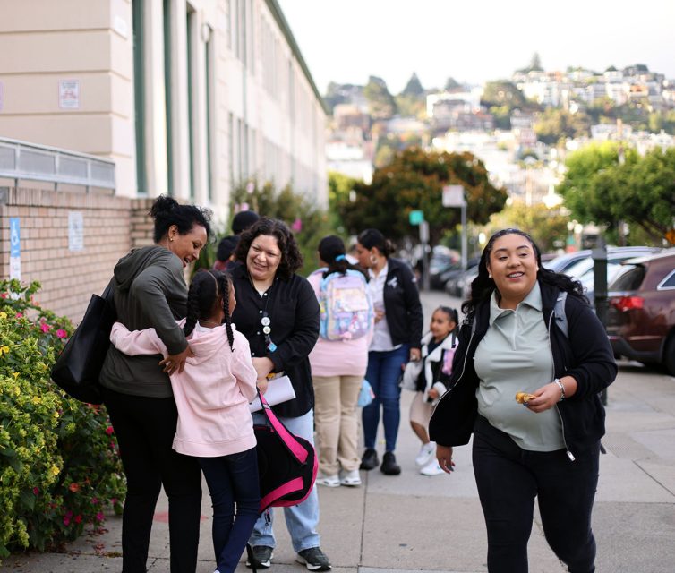 A group of parents, children, and a teacher conversing and hugging on a school sidewalk, with a hilly neighborhood in the background.
