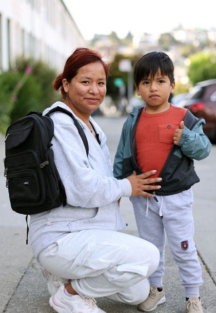 A woman with a backpack kneels next to a young boy giving a thumbs-up. Both are dressed in casual attire and are outdoors on a city street.