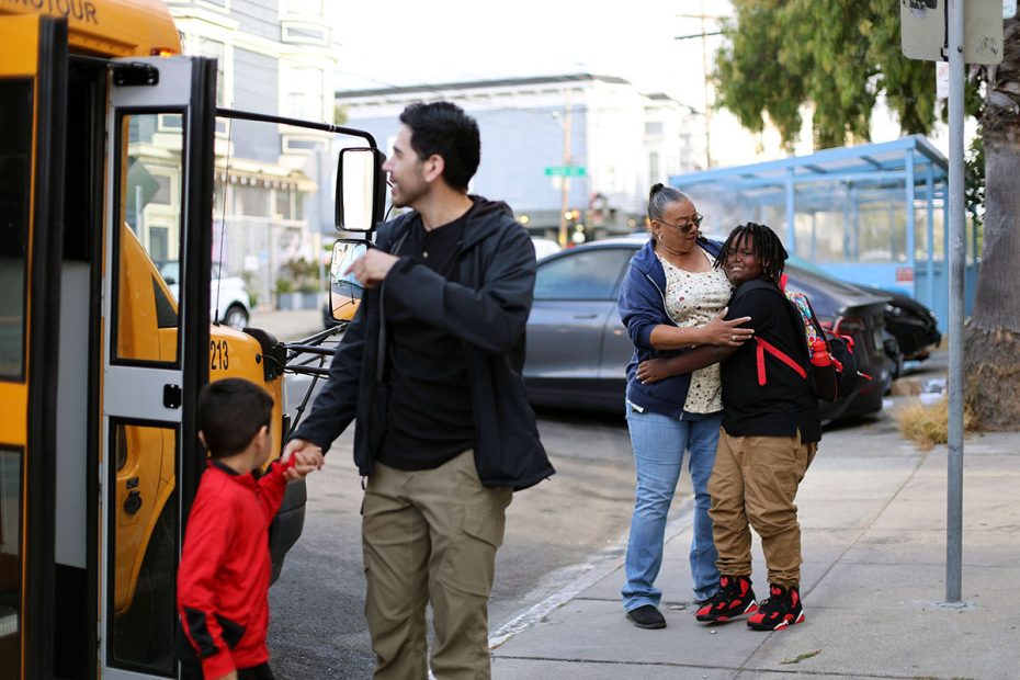 A man holds a child's hand while walking past a school bus. A woman hugs another child near a car on the sidewalk.