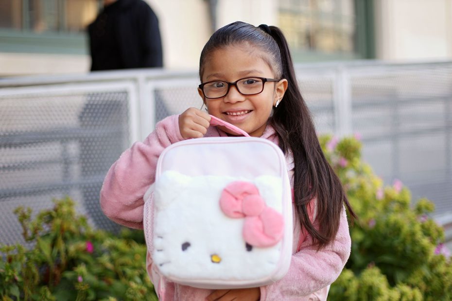 A young girl with glasses and a pink jacket smiles while holding a Hello Kitty lunch bag, standing outside in front of a metal railing.