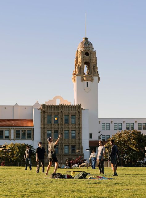 People playing bocce on a grassy field in front of a historic building with a bell tower.