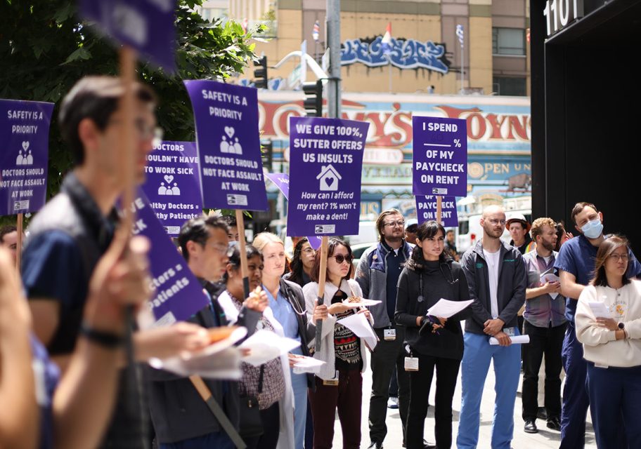 A group of people is gathered outdoors, holding signs with messages about workplace safety and fair compensation. Some signs read "Safety is a priority" and "How can I afford my rent if 2/3 of my paycheck goes?.