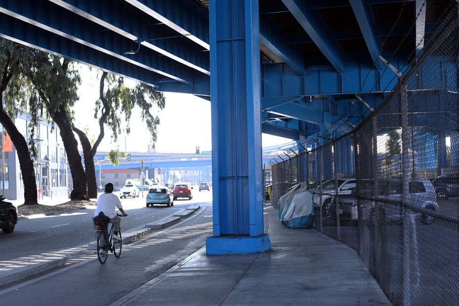 A person rides a bicycle under a blue-painted overpass with cars and parked vehicles along the sides and a fence to the right. Trees and another overpass are visible in the distance.
