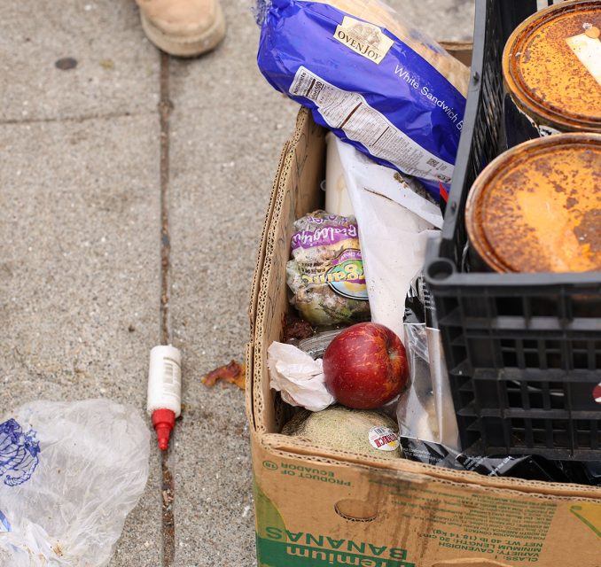 A cardboard box containing a variety of items including a loaf of bread, pomegranate, plastic containers, and a tube of adhesive sits on a concrete sidewalk.