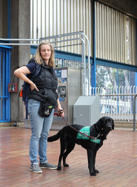 A woman stands on a tiled platform wearing a black vest and carrying a backpack, holding a leash attached to a black service dog wearing a green vest.