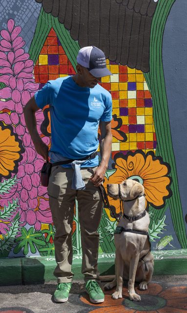 A man wearing a blue shirt, cap, and khaki pants stands next to a dog on a leash in front of a colorful mural with flowers.
