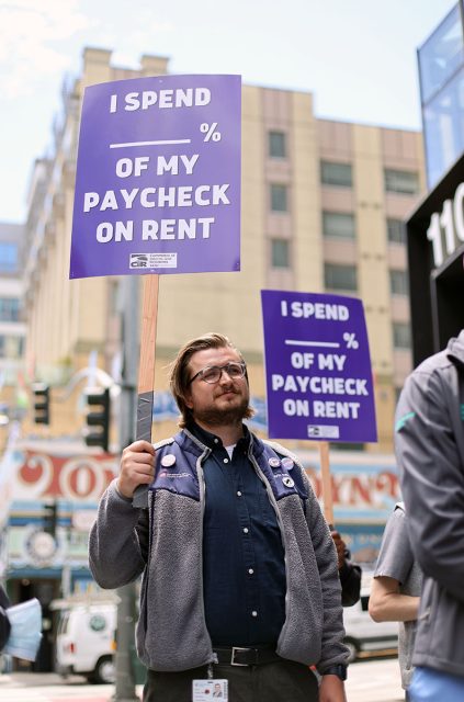 A person holds a purple sign that reads, "I spend % of my paycheck on rent," during a daytime protest. Another similar sign is visible in the background.