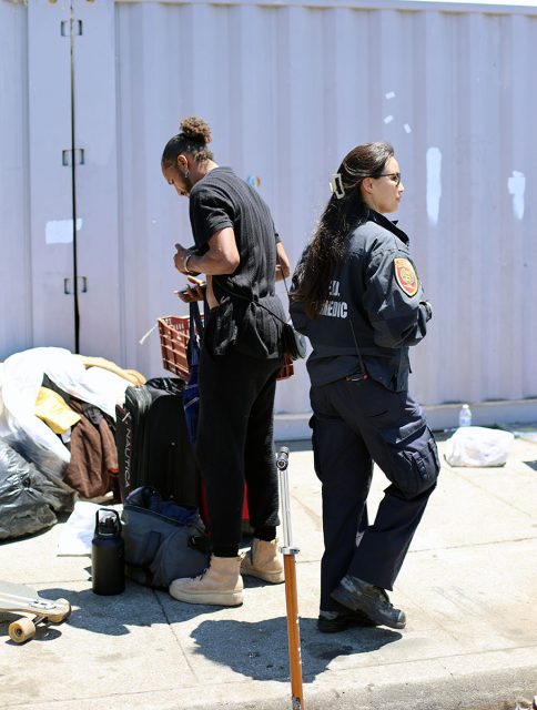 A person stands near packed belongings and a skateboard while another individual, dressed in a uniform, stands beside them on a street near a container.